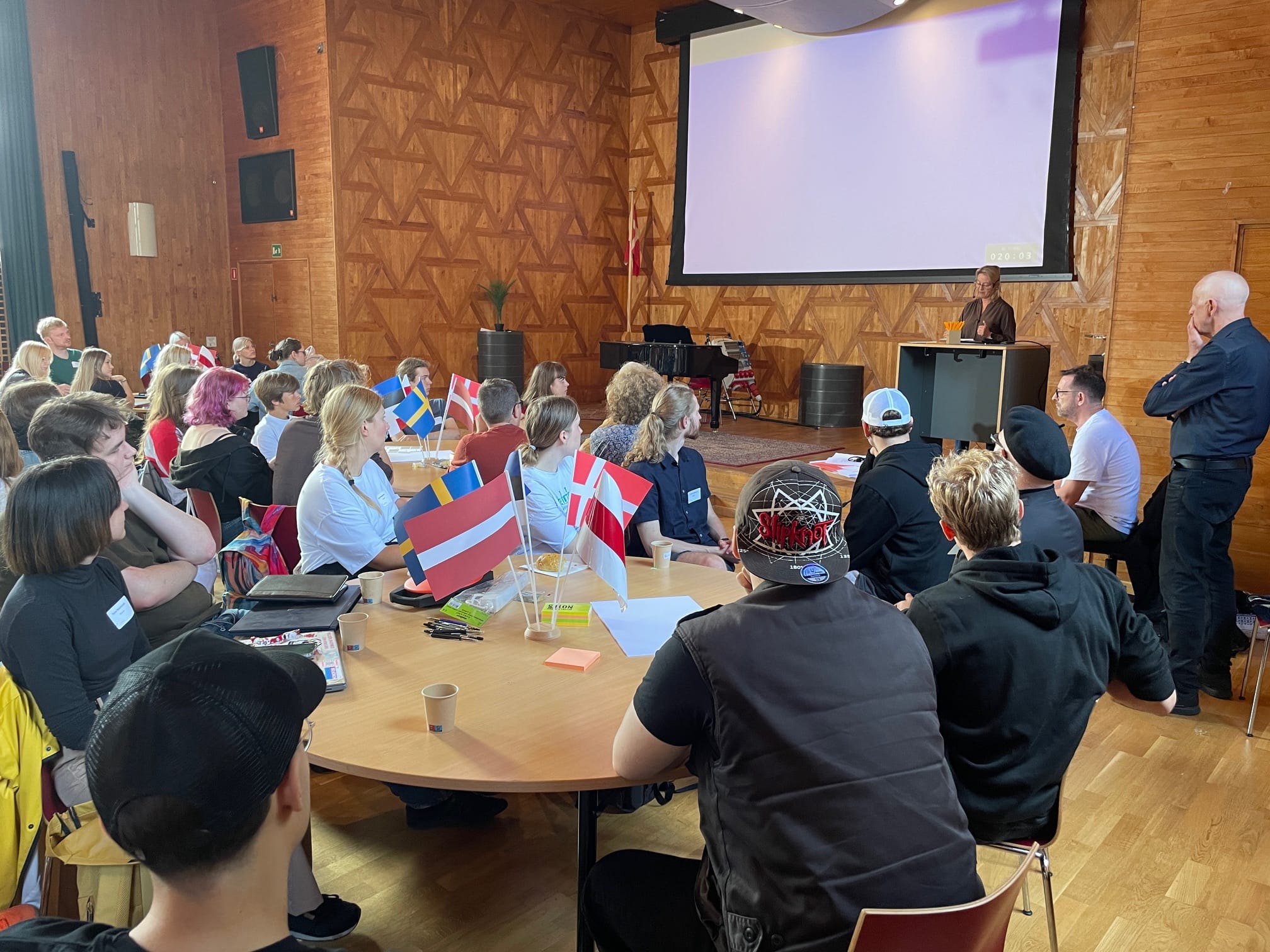En grupp människor sitter vid bordsgrupper i ett konferensrum och lyssnar till en talare som står vid en talarpult framför en stor skärm. Deltagarna representerar olika länder, vilket syns på de små flaggorna från Danmark, Sverige och andra nationer som är placerade på borden. Lokalen har en vägg med träpaneler och atmosfären verkar engagerad och fokuserad.