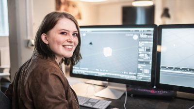 Tjej som sitter framför två datorskärmar och ler in i kameran.