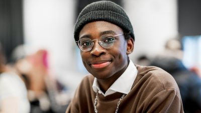 En elev på NTI Gymnasiet med mössa, glasögon och brun tröja tittar in i kameran och ler
