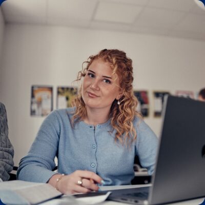 En elev med lockigt hår i klassrum vid dator och juridikbok som tittar in i kameran.