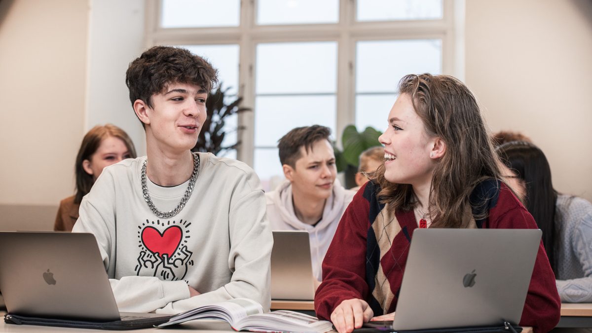 Flera elever sitter i klassrummet. Närmst kameran sitter en kille och en tjej vid sina datorer och pratar under en lektion