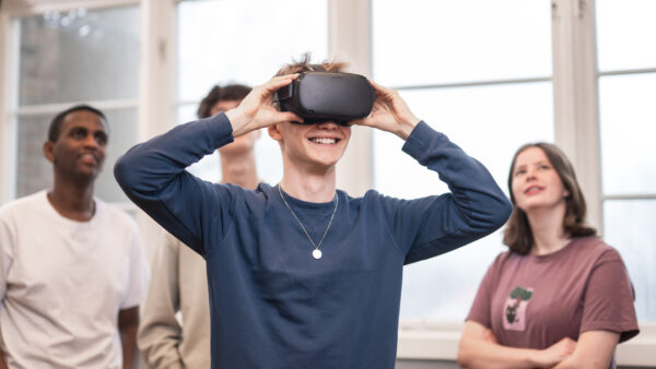 4 elever står och spela med VR. Killen längst fram har på sig VR-glasögon och de andra står bakom och tittar upp på en skärm som visar vad han ser.