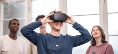 4 elever står och spela med VR. Killen längst fram har på sig VR-glasögon och de andra står bakom och tittar upp på en skärm som visar vad han ser.