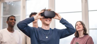 4 elever står och spela med VR. Killen längst fram har på sig VR-glasögon och de andra står bakom och tittar upp på en skärm som visar vad han ser.