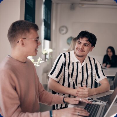 Två elever vid datorer i klassrummet som pratar med varandra.
