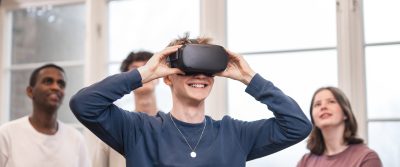 4 elever står och spela med VR. Killen längst fram har på sig VR-glasögon och de andra står bakom och tittar upp på en skärm som visar vad han ser.