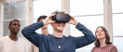 4 elever står och spela med VR. Killen längst fram har på sig VR-glasögon och de andra står bakom och tittar upp på en skärm som visar vad han ser.